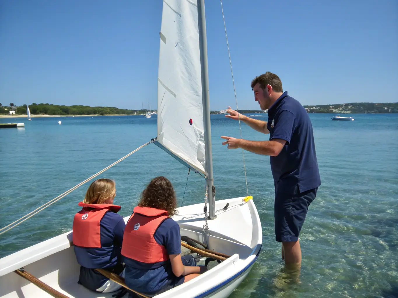 A group of children learning to sail small boats in a calm bay, with an instructor nearby providing guidance. The scene is bright and sunny, emphasizing the joy and accessibility of sailing.